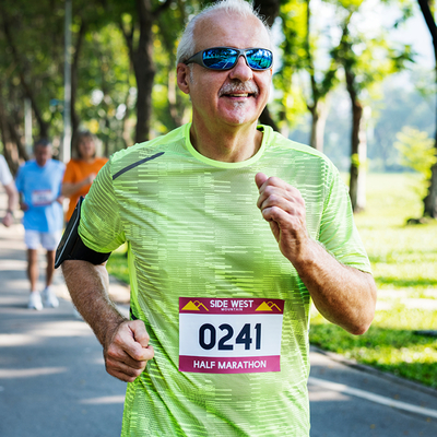 Un homme souriant d'âge moyen, portant un t-shirt vert fluo et des lunettes de soleil, court en plein air avec un dossard de marathon imprimé sur Polyart Laser Soft.