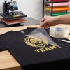 Main retirant le film de transfert transparent POLI-TACK 854 d'un t-shirt noir floqué d'un motif lion doré