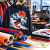 Sweat à capuche personnalisé avec motif skieur floqué au flex thermique POLI-FLEX TURBO à côté d'une presse à chaud et de rouleaux de couleurs